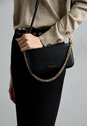 Black leather crossbody bag with a textured surface, gold chain strap, and branded detail. Paired with a beige top and black trousers.