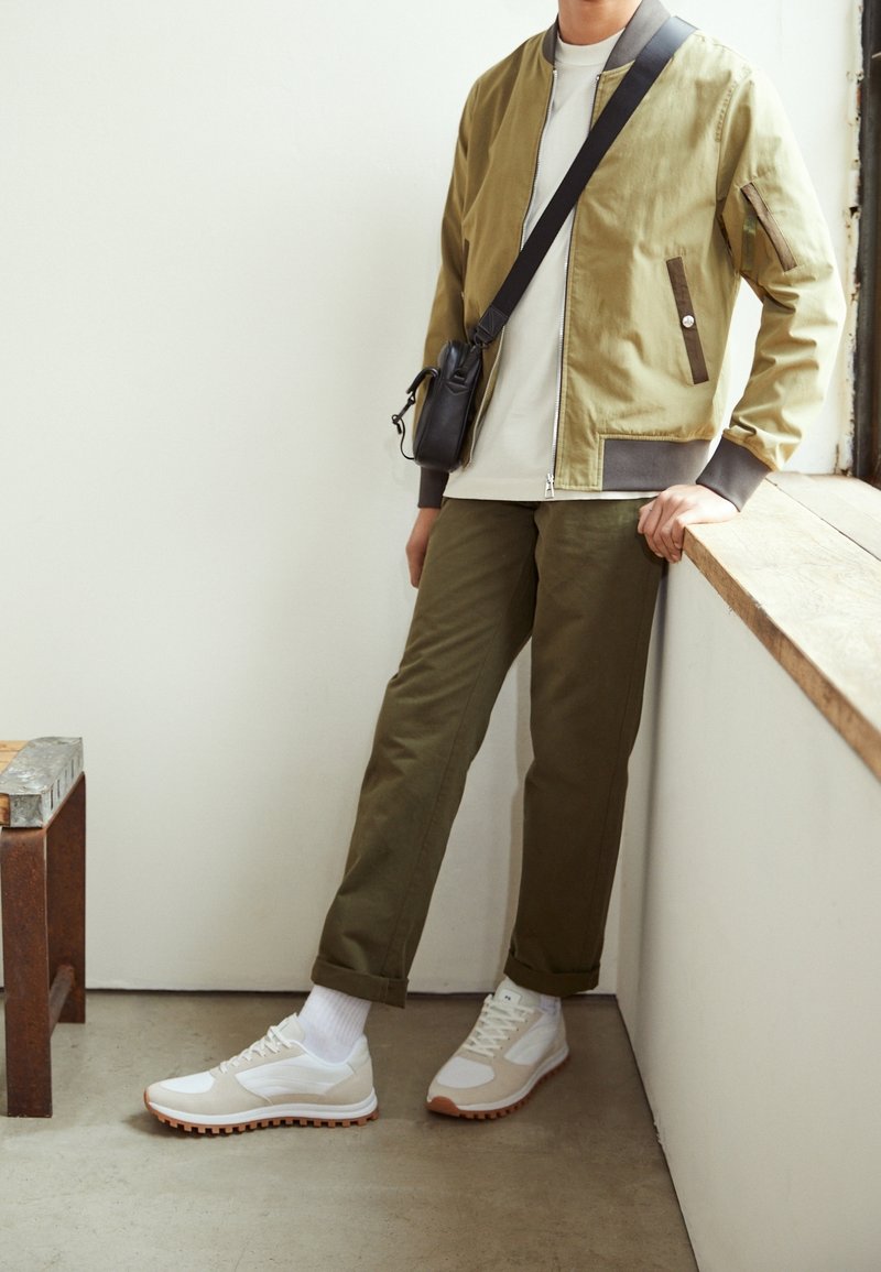 Personne portant une veste aviateur kaki, un pantalon olive, des baskets blanches et un sac bandoulière noir, appuyée contre un rebord en bois près d'une fenêtre.