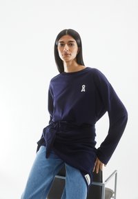 Navyblaues Langarmshirt mit einem Ripsband-Logo auf der Brust, kombiniert mit hellblauen Jeans. Der Stoff wirkt weich und hat eine lockere Passform.