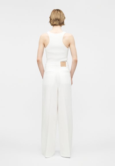 Άτομο με κοντά ανοιχτό καστανά μαλλιά, που φοράει λευκό αμάνικο ριμπ τοπ και λευκό παντελόνι με ψηλή μέση και φαρδιά μανίκια, από πίσω.