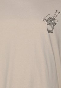 Beige T-shirt featuring a printed graphic of a takeout box with noodles and chopsticks, labelled "YOKOHAMA CHINATOWN." Soft cotton texture.