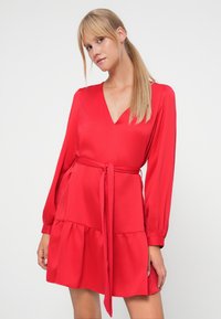 Robe rouge avec un décolleté en V, des manches longues et une taille ceinturée. Elle présente un ourlet à volants et une texture de tissu lisse.