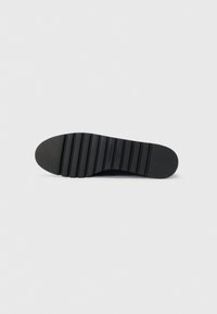 Semelle de chaussure en caoutchouc noir avec un motif texturé et nervuré. Elle présente une forme arrondie et un détail de logo en relief près du centre.