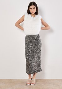 Top senza maniche in pelliccia sintetica bianca abbinato a una gonna midi argentata con paillettes. La modella indossa sandali bianchi con cinturini, mostrando un outfit strutturato.