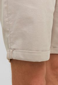 Pantalones cortos beige hechos de una tela texturizada, con un dobladillo enrollado y detalles de costura sutiles. El diseño destaca un aspecto limpio y entallado.