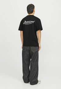 Camiseta de algodón negra con cuello redondo y mangas cortas. Presenta un texto de logo en blanco en la parte posterior. Combinada con unos jeans de mezclilla oscuros y holgados.