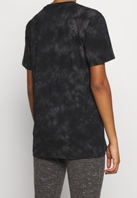 Persona vestida con una camiseta de manga corta negra y con diseño de tie-dye y pantalones grises texturizados, mostrada desde atrás sobre un fondo simple.