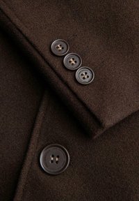 Tumma ruskea villakangastakki lähikuvassa, jossa näkyy neljä tummaa ruskeaa nappia ja teksti, korostaen kankaan rakennetta ja nappien yksityiskohtia.