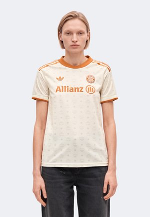 Un mannequin porte un maillot de football crème du Bayern Munich, avec des finitions orange et des logos de sponsors, associé à un jean noir sur un fond clair uni.