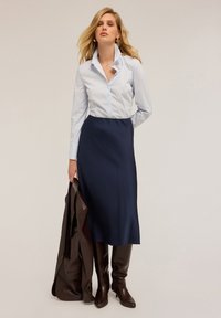 Camicia a righe azzurro chiaro con colletto, abbinata a una gonna blu scuro aderente. Un giubbotto di pelle marrone e stivali marroni fino al ginocchio completano il look.