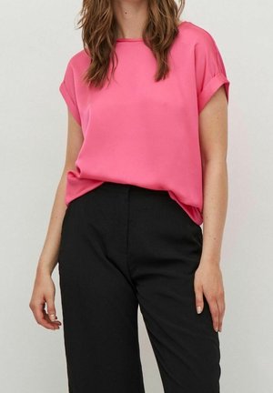 Frau trägt eine knallpinke kurzärmelige Bluse, die in eine hoch taillierte schwarze Hose gesteckt ist, steht mit locker hängenden Armen an den Seiten.