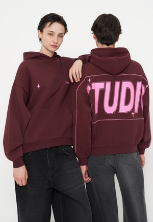 Zwei Personen tragen weinrote Kapuzenpullover mit pinkem Text und Stern-Designs, eine blickt nach vorne, die andere zeigt großen "STUDI"-Text auf dem Rücken.