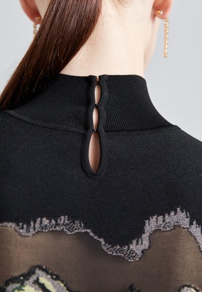Black high-neck garment with a textured knit and sheer panel featuring floral design. Back keyhole closure accentuated with small loops.