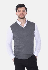 Gilet grigio lavorato a maglia con scollo a V, indossato sopra una camicia bianca con bottoni. Presenta orlo e aperture delle maniche ribattuti.