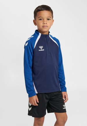 Dreng iført en blå og mørkeblå sportstop med kvart lynlås og sorte shorts med hvidt logo og chevron-detaljer står med fronten mod fronten.