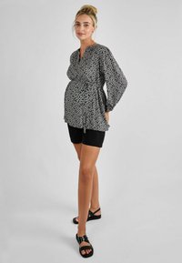 Schwarz-weiße Bluse mit Polka-Dots, langen Ärmeln, V-Ausschnitt und lockerer Passform. Kombiniert mit schwarzen Shorts und flachen Sandalen.