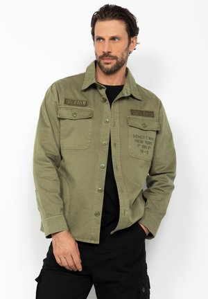 Mężczyzna w oliwkowo zielonej koszuli zapinanej na guziki z kieszeniami na piersi i naszywkami, w czarnych spodniach, stoi na tle białej ściany.