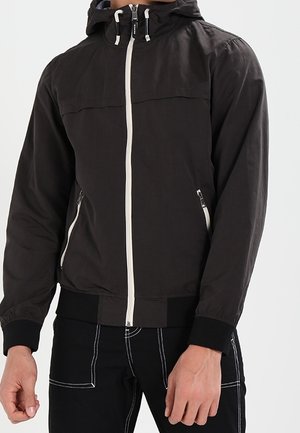 Chaqueta con capucha negra con cremallera blanca y cremalleras en los bolsillos, llevada por una persona vestida con pantalones negros con costuras blancas.