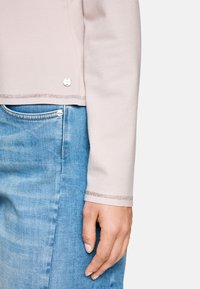 Helles rosa Oberteil mit langen Ärmeln und silberner Borte an den Ärmelabschlüssen, kombiniert mit hellblauen Jeans, die mit Metallknöpfen verziert und in einer ausgewaschenen Optik gehalten sind.