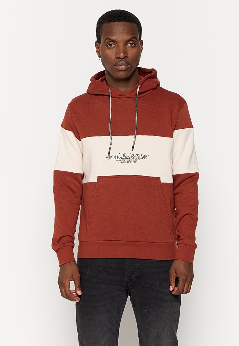Homme portant un hoodie coloré en rouille et blanc avec une poche avant et un jean noir, se tenant devant un fond blanc uni.