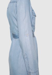 Ljusblå denimklänning med figursydd midja, knappdetaljer, långa ärmar och bröstfickor. Slät textur, klassisk design.