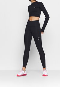 Schwarzes langärmliges Cropped-Top und hochtaillierte Leggings aus dehnbarem Material. Weißes Logo auf dem Top und den Leggings. Schuhe: weiß mit pinken Akzenten.