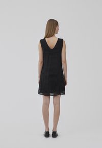 Robe noire sans manches avec un dos en V, présentant un motif à carreaux texturé et une coupe ample, tombant au-dessus des genoux. Des chaussures noires complètent le look.