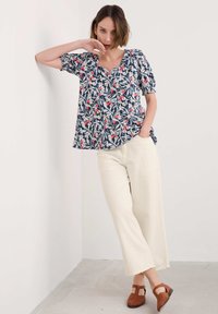 Kratkām piedurknēm, tumši zila ziedu blūze ar baltām un sarkanām apdrukām, apvienota ar krēmkrāsas platām biksēm un brūnām austām sandālēm.