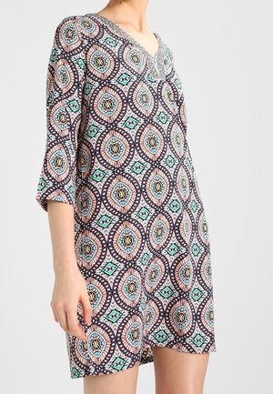 Kobieta w luźnej sukience do kolan z rękawami 3/4 i wielokolorowym geometrycznym wzorem w odcieniach niebieskiego, różowego i czarnego.