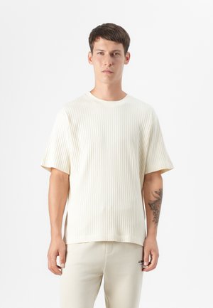 Een jonge man staat tegen een effen achtergrond, gekleed in een crèmekleurig geribbelde t-shirt en lichtbeige broek, en toont een tattoo op zijn arm.