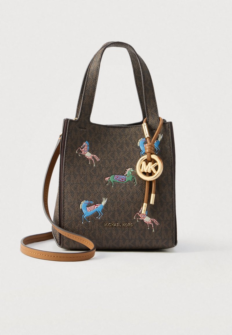 Bruine Michael Kors-handtas met korte handvatten, paardenapplicaties, gouden MK-bedel en lange afneembare crossbodyriem tegen een effen achtergrond.