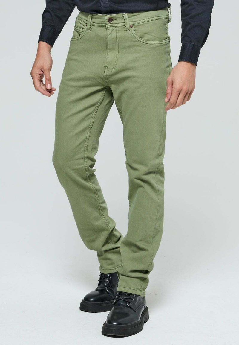 Jean en mélange de coton vert avec une coupe slim, comportant des poches avant, une fermeture à bouton et des détails de surpiqûres visibles. Porté avec des bottes noires.