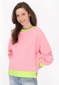 Felpa rosa con vestibilità oversize, colletto e polsini in festone verde, morbido tessuto di cotone, spacchi laterali all'orlo, abbinata a jeans azzurri.
