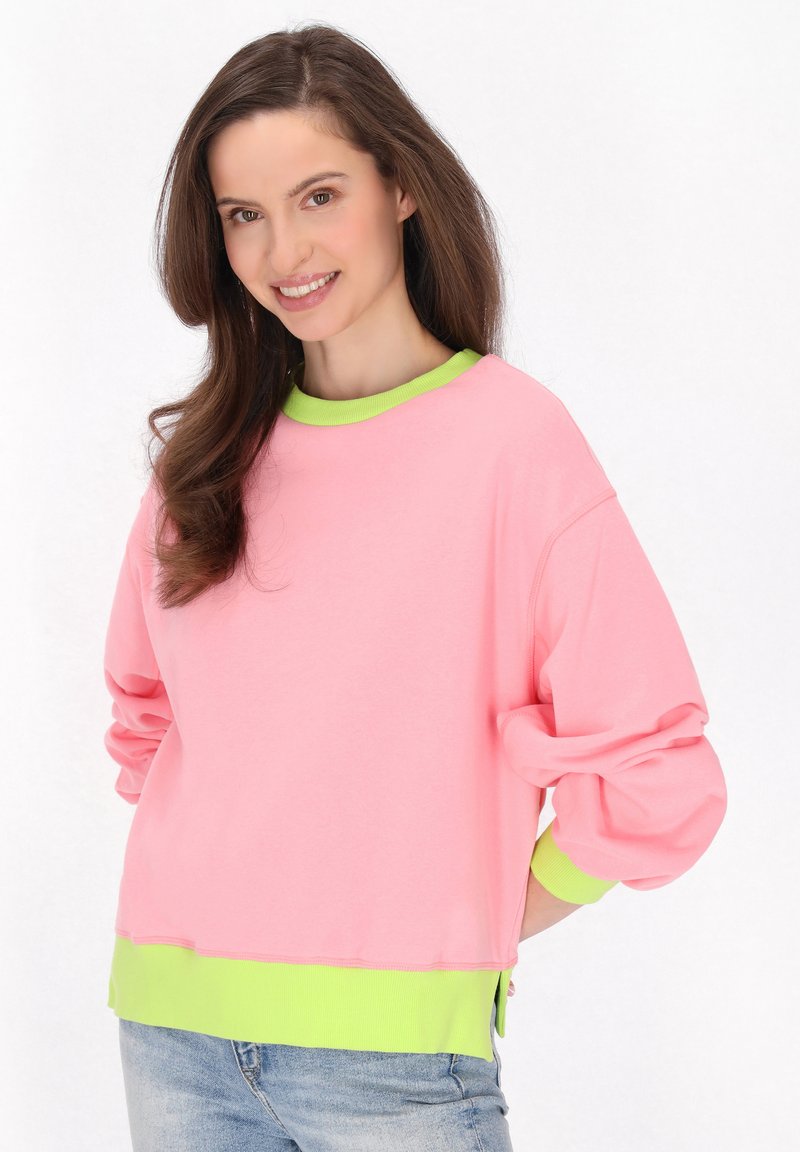Felpa rosa con vestibilità oversize, colletto e polsini in festone verde, morbido tessuto di cotone, spacchi laterali all'orlo, abbinata a jeans azzurri.