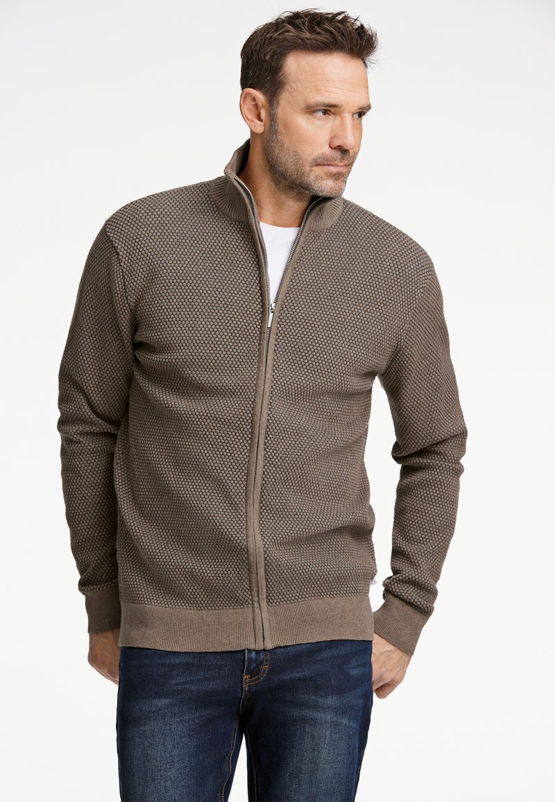 Brun zip-up tröja med ett texturerat bikakemönster och hög krage. Har ribbstickade muddar och fåll, kombinerad med blå jeans.