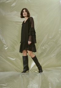 Svart, lager-på-lager klänning med genomskinliga volangärmar och fåll; kombinerad med knähöga svarta klackstövlar; mot en bakgrund av lätt plast.