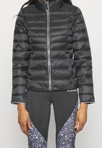 Vrouw die een zwart gewatteerd jack draagt met een voorrits en zwarte legging met grijze en zwarte luipaardprintpanelen.