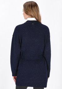 Marinblå stickad kofta med långa ärmar, bälte i midjan, ribbad nederkant och prickiga texturdetaill. Har en rund halsringning och avslappnad passform.