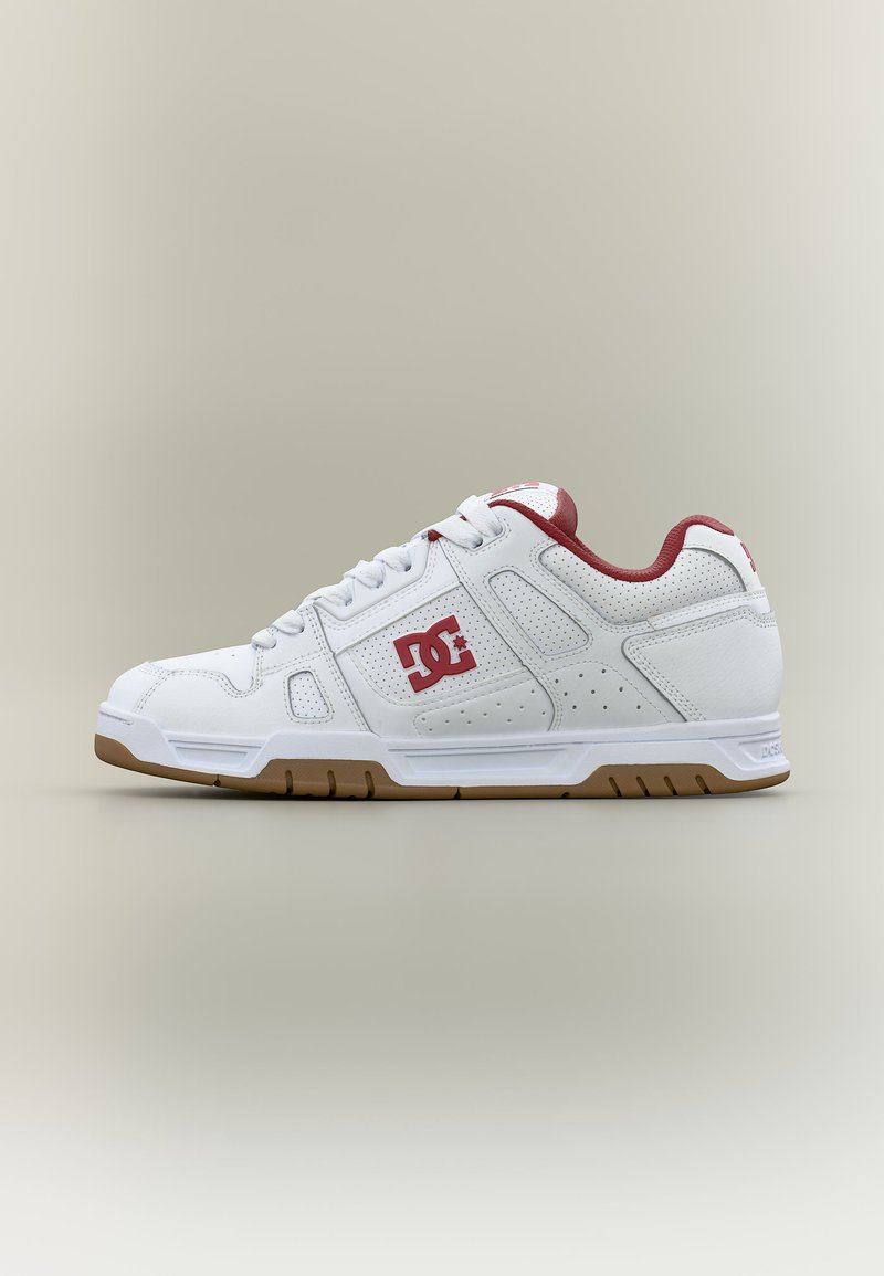 Baskets blanches basses avec doublure intérieure rouge, logo DC sur le côté, panneaux perforés et semelle couleur gomme sur un fond neutre.