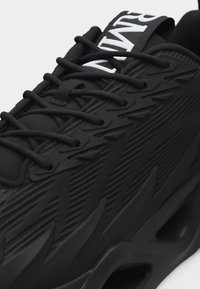 Μαύρο αθλητικό παπούτσι με υφή σχεδίου, μαύρα κορδόνια και έντονο λευκό λογότυπο «NIKE» στη γλώσσα.