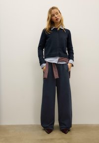 Dunkelblauer Cardigan mit Knopfleiste, darüber ein gestreiftes Hemd getragen. Weite marineblaue Hosen mit einem rot-weiß gestreiften Gürtel und spitze bordeauxrote Schuhe.