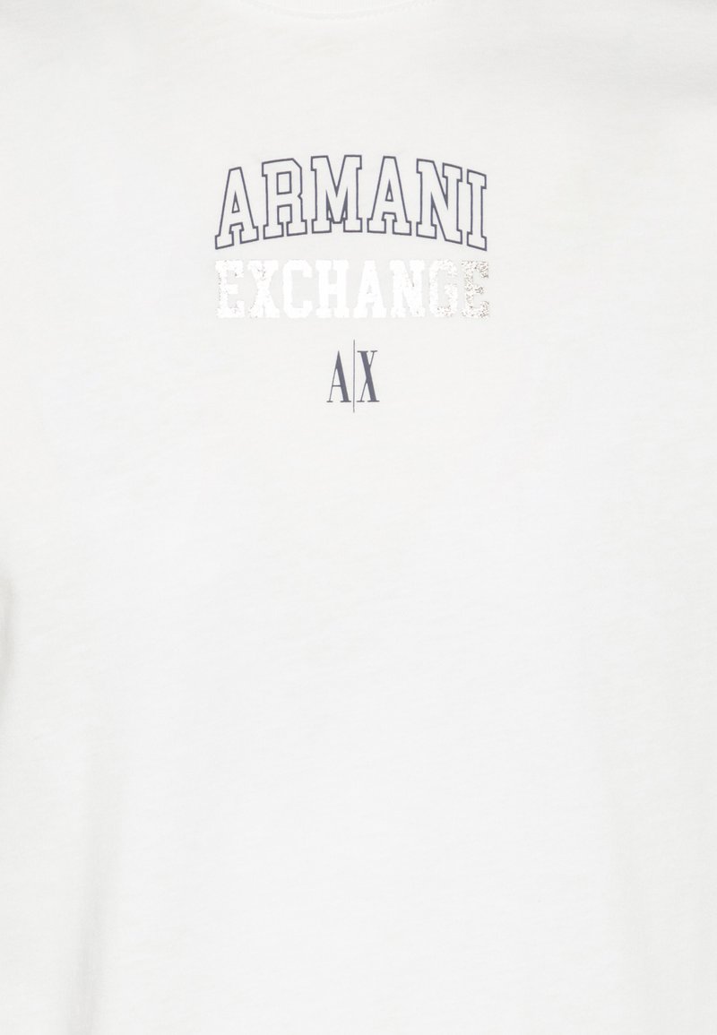 Camiseta blanca con el logo de "Armani Exchange" en gris, con una textura desgastada. Diseño sencillo, de mangas cortas y con cuello redondo.