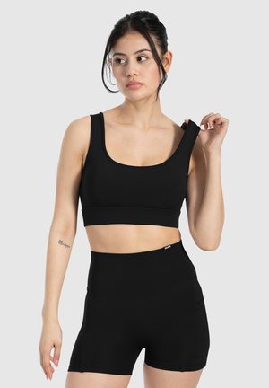 Schwarzes Sport-Crop-Top mit breiten Trägern und quadratischem Ausschnitt, kombiniert mit hochgeschnittenen schwarzen Shorts mit Seitentaschen. Weicher, dehnbarer Stoff.
