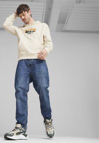 Felpa crema con logo "Puma", tasca frontale, abbinata a jeans blu larghi e sneakers chunky grigio-verdi.