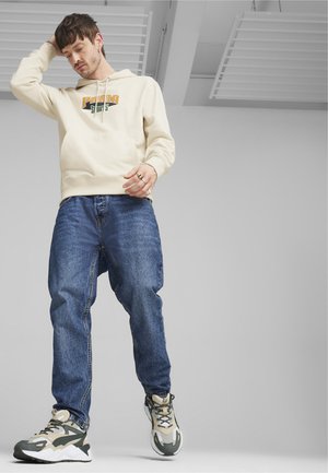 Felpa crema con logo "Puma", tasca frontale, abbinata a jeans blu larghi e sneakers chunky grigio-verdi.
