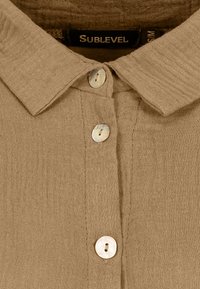 Beige Leinenhemd mit Kragenaufbau, ausgestattet mit drei runden, hellen Knöpfen und einem strukturierten Stoff. Markenlabel sichtbar.