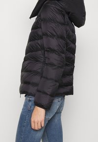 Sort pufferjakke med hætte, der har et quiltet design, elastiske manchetter og en lynlås. Bæres over blå jeans.