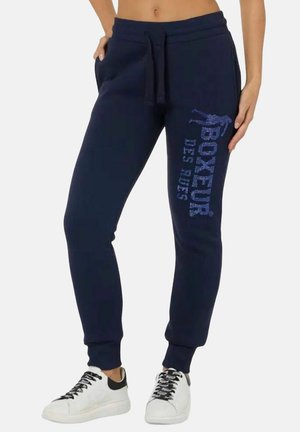 Pantaloni jogger navy con vita regolabile e testo "Boxeur Des Rues" e grafica di boxer sulla coscia sinistra, indossati con sneaker bianche.