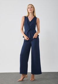 Top peplum sans manches bleu marine avec décolleté en V et détail torsadé à la taille, associé à un pantalon large assorti bleu marine et des talons nus.