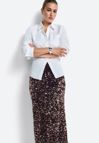 Fehér gombos ing egy figyelemfelkeltő, hosszú, sötét színű flitteres szoknyával párosítva. Az ezüst karkötő kiemeli a csuklót. A sima textúra kontrasztot képez a flitterekkel.